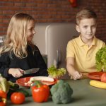 Positive little girl and boy sitting at table with assorted vegetables and fruits while preparing healthy food together in loft kitchen with brick wall